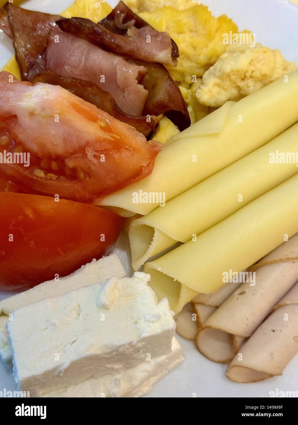 Frühstücksteller mit Rührei, Speck, Schinken, Käse, frischen Tomaten, Feta-Käse und Feinkost-truthahn. - Smartphone-aufgenommenes Stockfoto