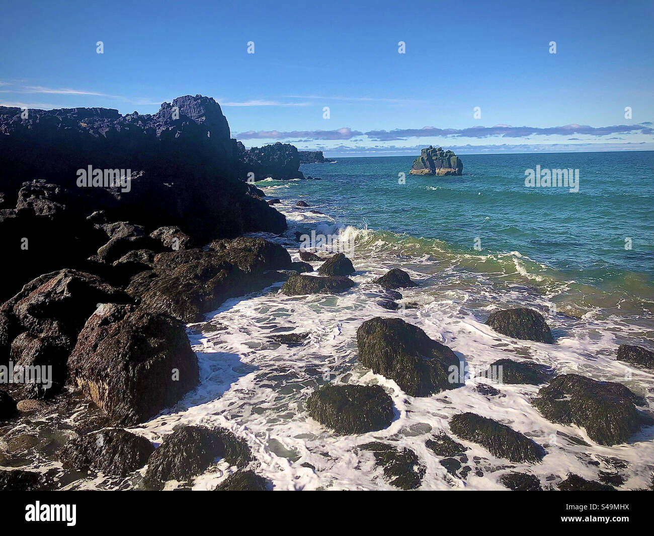 Hohe Klippe mit großen Felsen im Wasser an der Küste in Island. Wellen krachen an Land und Blasen auf dem Wasser. Kleine Felseninsel in der Ferne. Himmelblau mit Wolken. Atlantik mit großen Wellen. Stockfoto