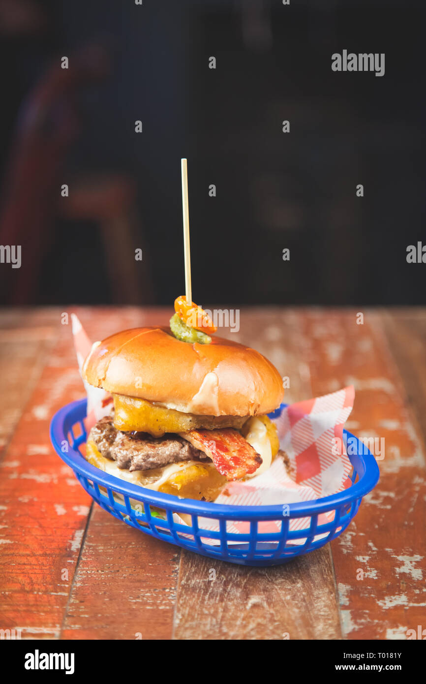 Beef Burger in Brioche Brötchen mit geschmolzenem Käse, Speck, Zwiebel Ringe serviert in einem Korb auf einem hölzernen Tisch in der Bar Stockfoto