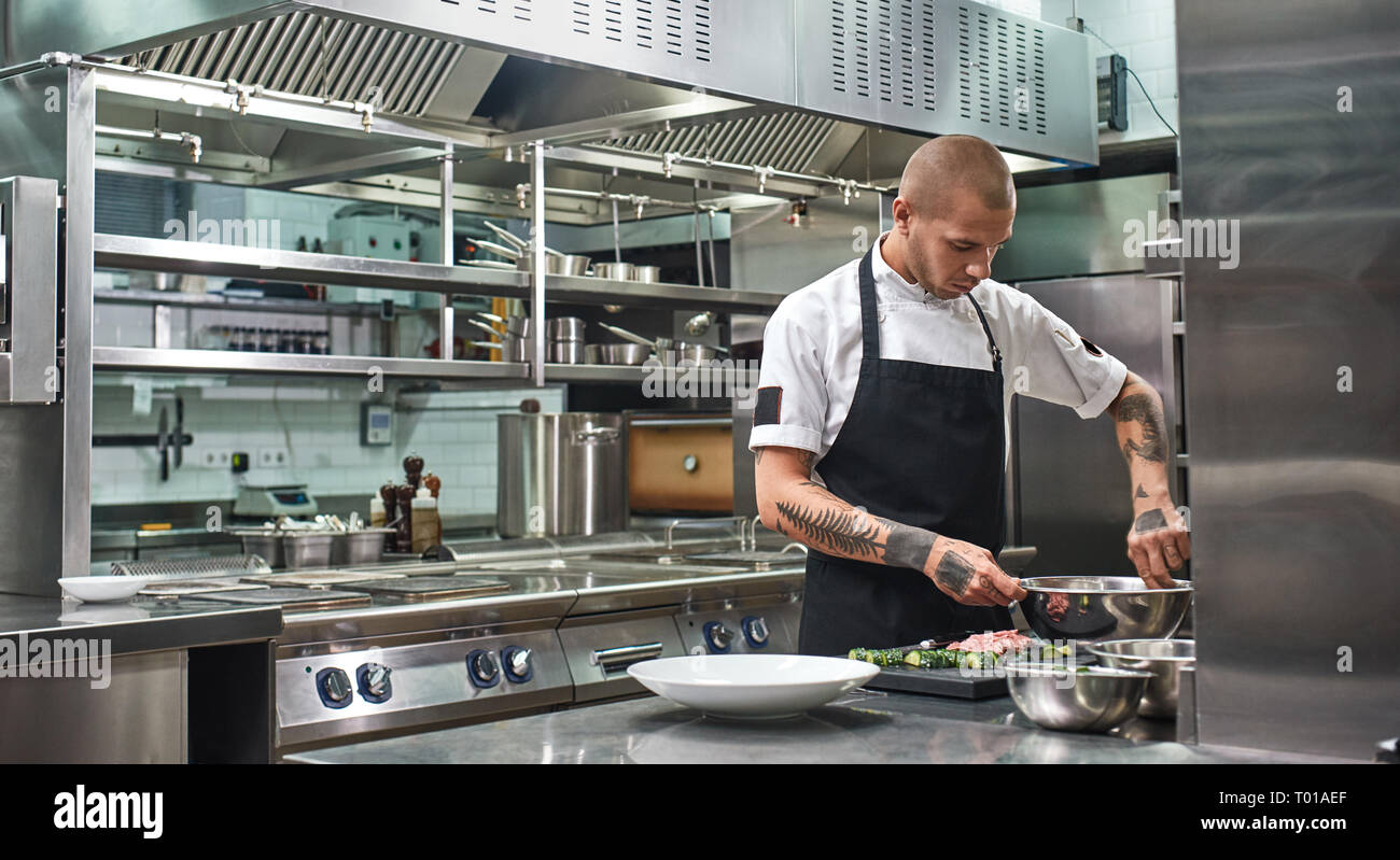 Fast fertig. Stattliche professioneller Koch mit Tattoos auf seinem Arme mischen einen Salat in einer Metallschüssel, während in der Küche stehen. Kochen Stockfoto