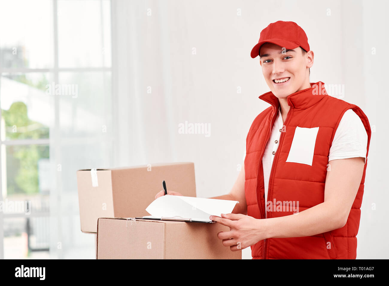 Porträt der Mann in der roten Uniform der Post in der Nähe von Stapel von Kisten, Studieren der Liste der Clients für heute. Freundliche Mitarbeiter, hohe Qualität. Im Innenbereich. Stockfoto