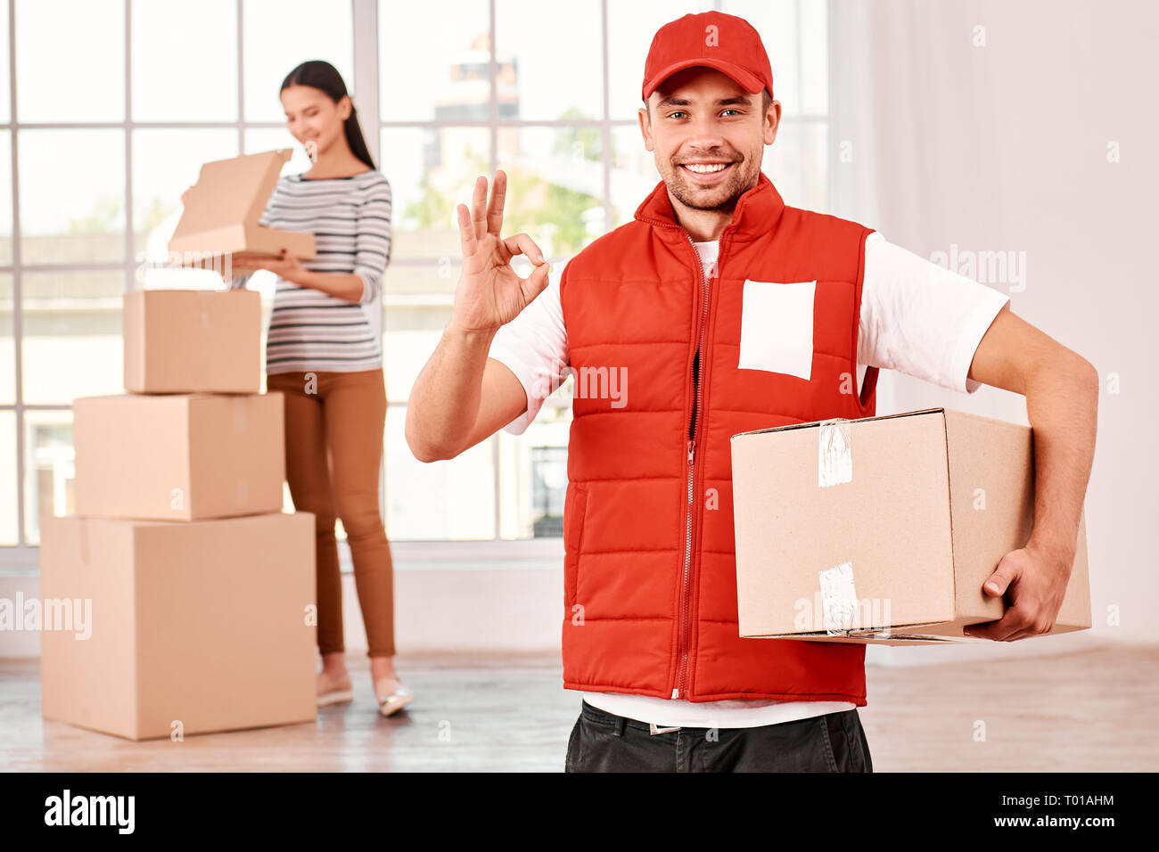 Junge Kurier in rot Post einheitliche stehend mit Paket drinnen, Anzeigen OK Zeichen, an der Kamera mit einem Lächeln. Er hat seinen Job sehr gut gemacht. Frau auspacken ihrer Einkäufe in den Hintergrund. Ein Stapel von Kartons. Hellen Innenraum. Freundliche Mitarbeiter, qualitativ hochwertige Service Stockfoto