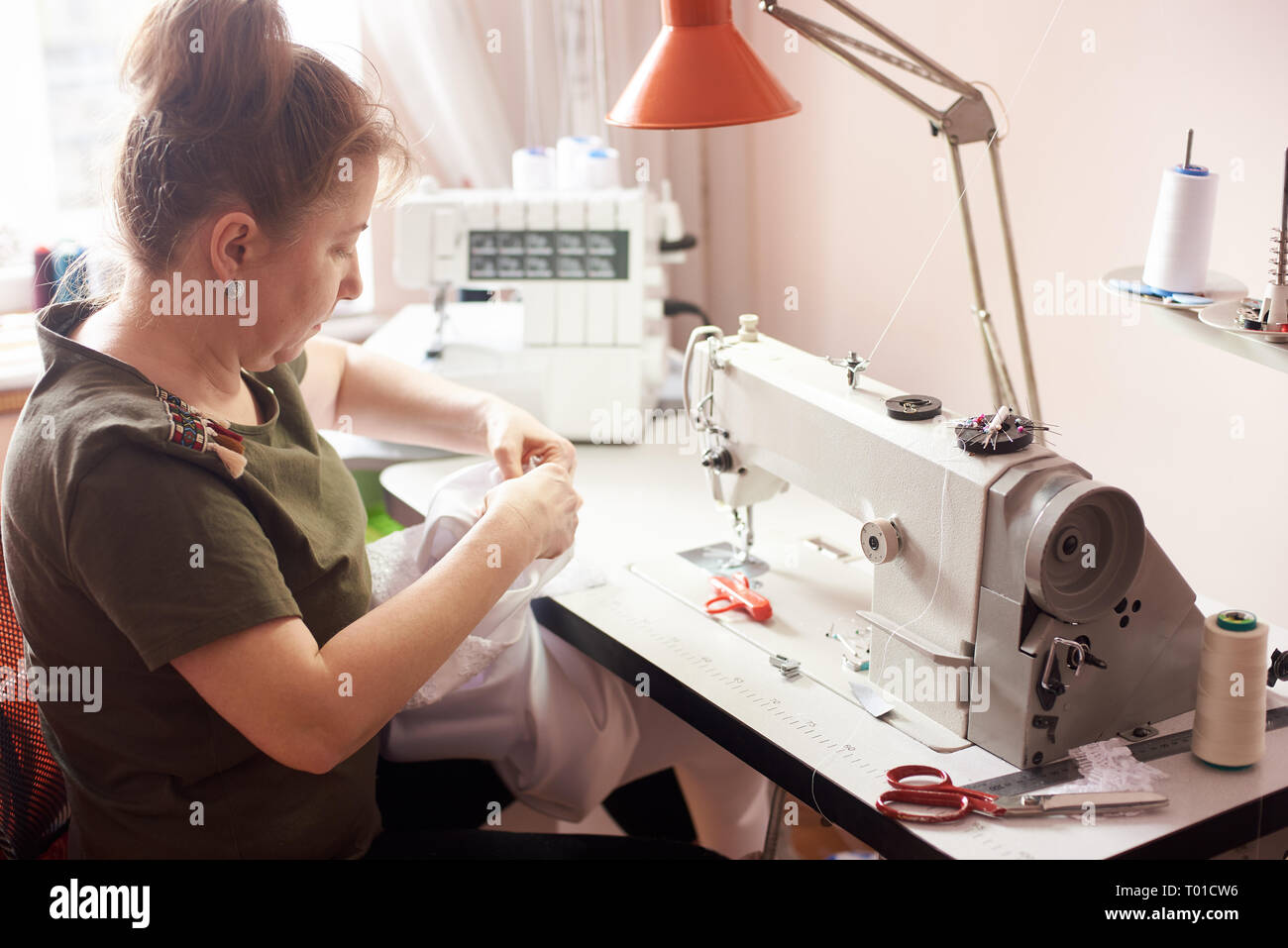 Textilhändler sitzen im Atelier, Stitching weißes Kleid mit Händen und Nadel. Näherin Arbeitsplatz mit Nähmaschine, Overlock, Schere, Stifte, Gewinde, Stockfoto