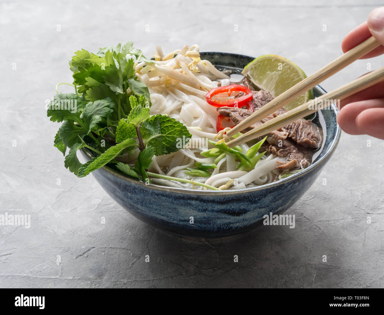 Traditionelle vietnamesische Suppe pho in eine blaue Schale, serviert mit Minze und cilantro, grüne Zwiebeln, Chili, Sojasprossen und Kalk. Hand mit einem Essstäbchen Stockfoto