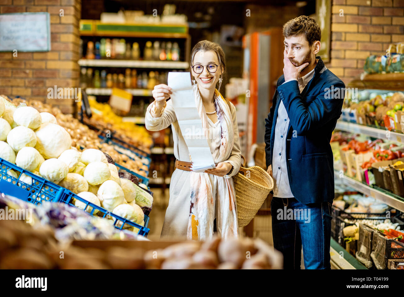 Junges Paar kaufen Essen gemeinsam mit langen Einkaufsliste in der Gemüseabteilung im Supermarkt. Mann mit schockiert Emotionen Stockfoto