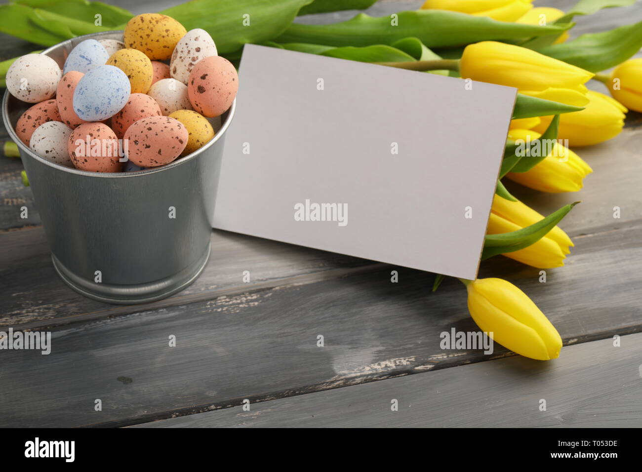 Bunte Ostern Süßigkeiten Eier mit gelben Tulpen und leere Grußkarte auf hölzernen Tisch Stockfoto