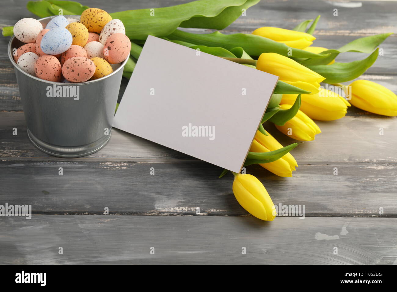 Bunte Ostern Süßigkeiten Eier mit gelben Tulpen und leere Grußkarte auf hölzernen Tisch Stockfoto
