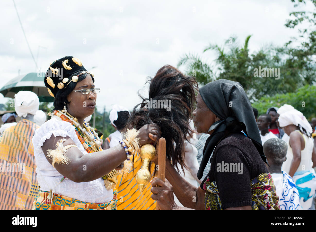 Adzopé, Elfenbeinküste - 31. August 2016: junge Frau in traditioneller Kleidung Halskette bells Hals gekleidet, Brille, Hut und Zopf in der Hand stehen Wi Stockfoto