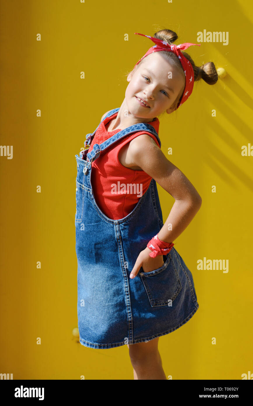 Portrait von niedlichen kleinen Mädchen in Jeans Anzug wearingred Weste und Kopftuch mit schwanz Haar. Das Kind lächelt, in der Nähe des gelben wand Stockfoto