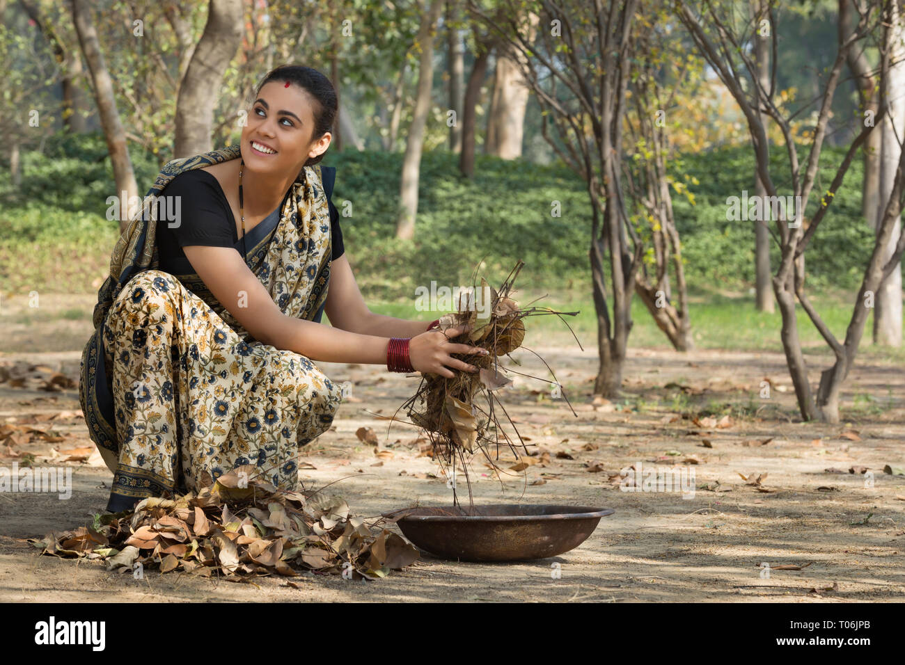 Lächelnd ländliche Frau, gekleidet in Saree sammeln getrocknete Blätter vom Boden in einem Eisen Gold Pan. Stockfoto