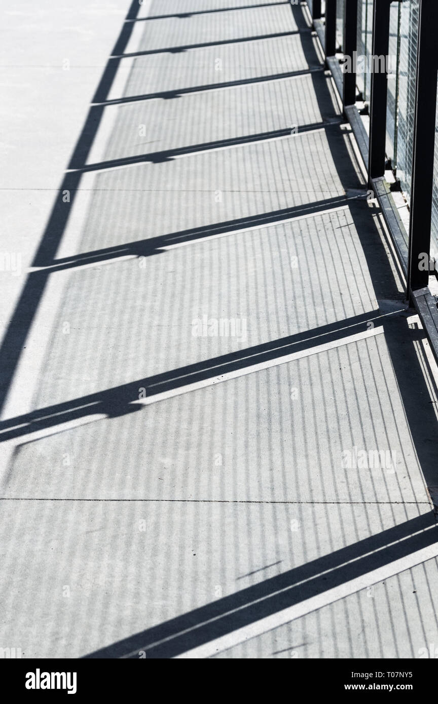Diagonale Schatten und Drahtgeflecht Muster auf Zement Gehweg aus Metall und Glas panel Zaun Barriere. Stockfoto