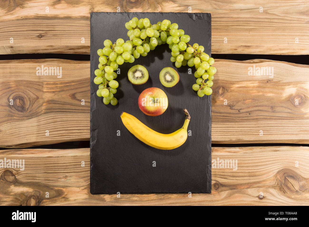 Glücklich essen - lächelndes Gesicht aus frischen Früchten flach zu legen. Schiefer server/Board/Fach auf rustikale Holzplanken Hintergrund. Banane, Apfel, Kiwi, Trauben. Stockfoto