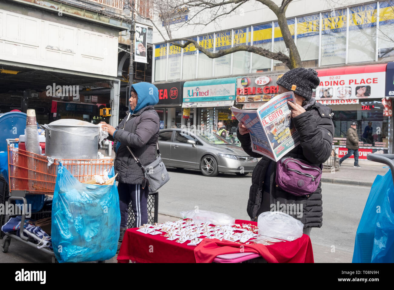 Eine Straße Szene auf 82nd St. in Jackson Heights, Queens, einem Anbieter liest eine spanische Zeitung und anderen bereitet Sie Lebensmittel zum Verkauf. Stockfoto
