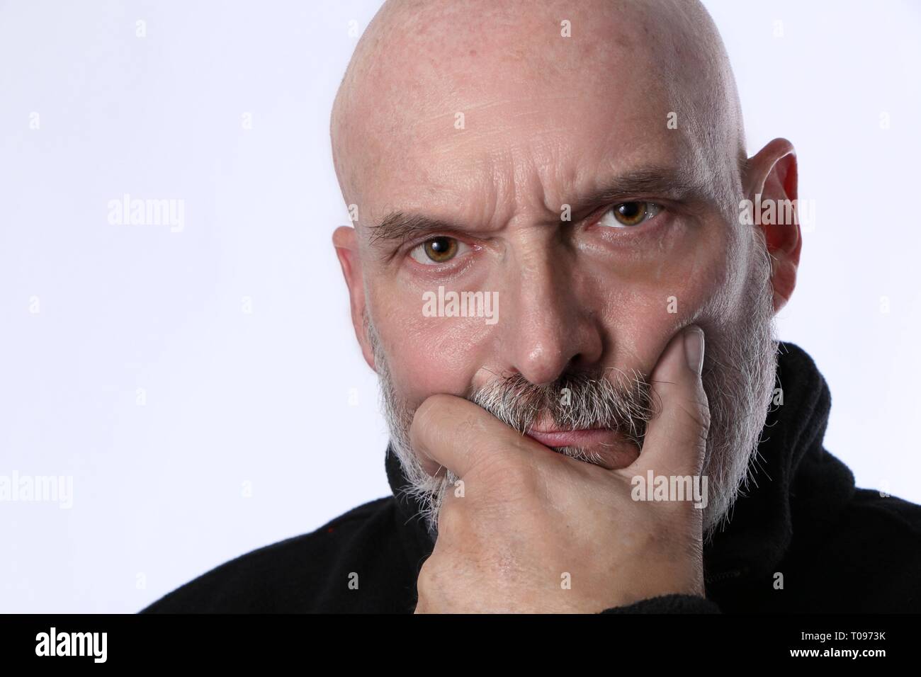 Älterer, Reifer Mann, Nachdenkliches Denken. Glatzkopf im Rentenalter mit der Hand im Gesicht. Durchstechend braune Augen auf weißem Hintergrund mit schwarzem Pullover. Stockfoto