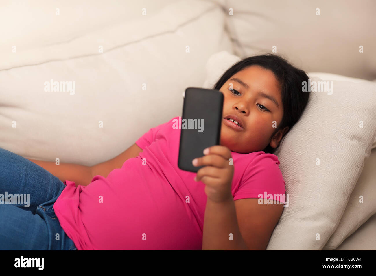 Niedliche latino girl Festlegung auf ein gemütliches Sofa mit einem Handy mit Freunden zu kommunizieren. Stockfoto