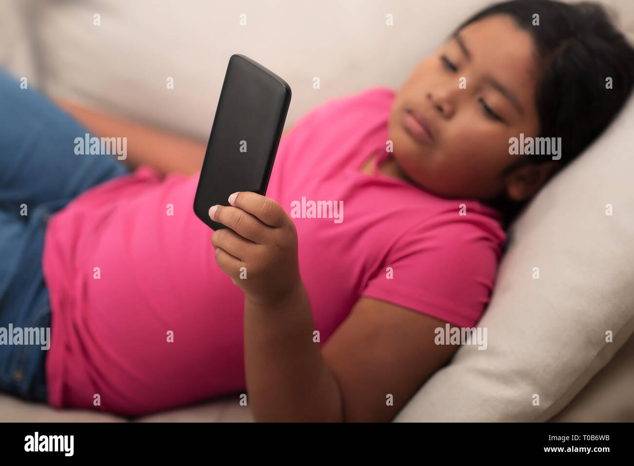 Hispanische Mädchen mit einem traurigen Blick auf ihrem Gesicht an einem Handy unbeaufsichtigt auf der Suche; ruht auf bequeme Couch. Stockfoto