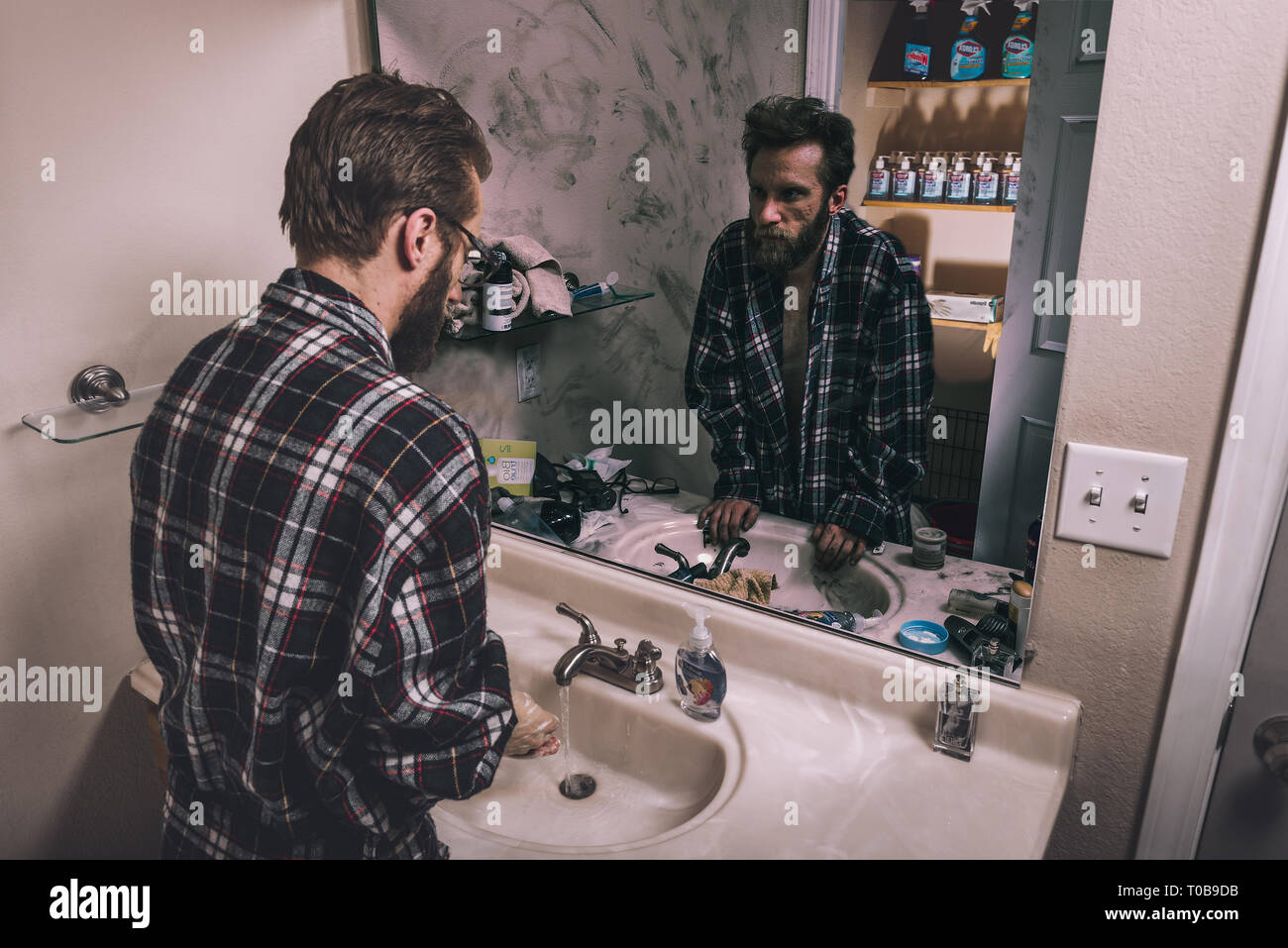 Ein junger Mann wäscht seine Hände in ein sauberes Bad, während sein ungepflegtes Reflexion auf sich selbst schaut, während in einem dreckigen Badezimmer stehen. Stockfoto