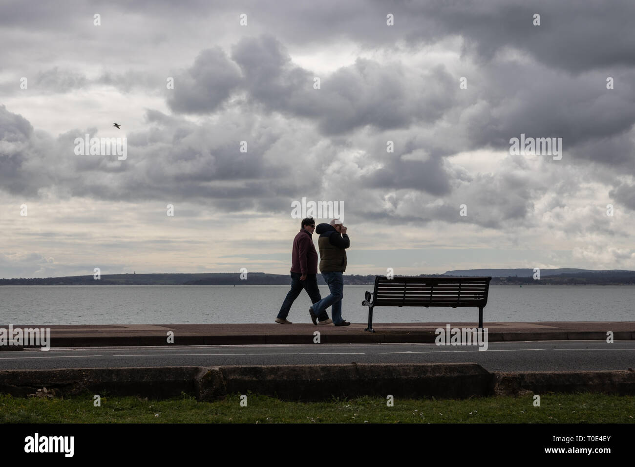 Ein Paar mittleren Alters zu Fuß entlang der Promenade am Strand an einem kalten und windigen Tag März Stockfoto