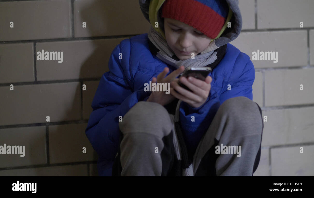 Junge sitzt auf dem Boden auf der Straße in den Abend und verwendet das Telefon im Winter Stockfoto