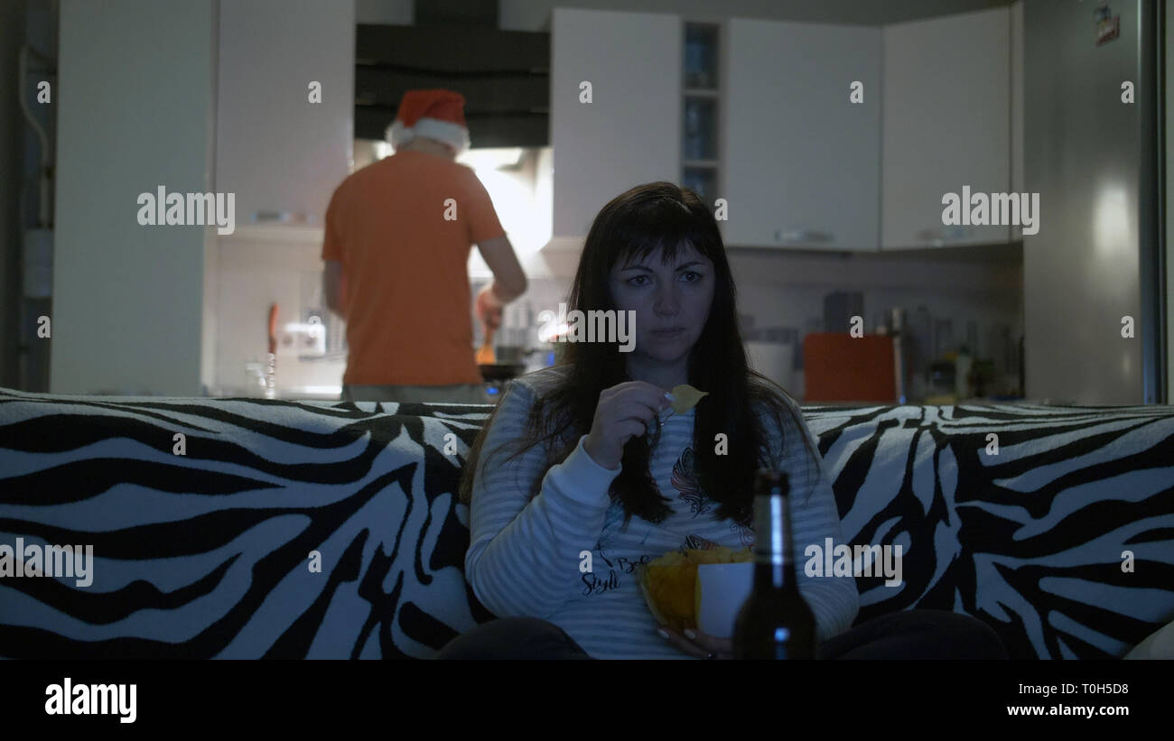 Frau vor dem Fernseher, trinkt Bier mit Chips, ein mann Essen bereitet Stockfoto