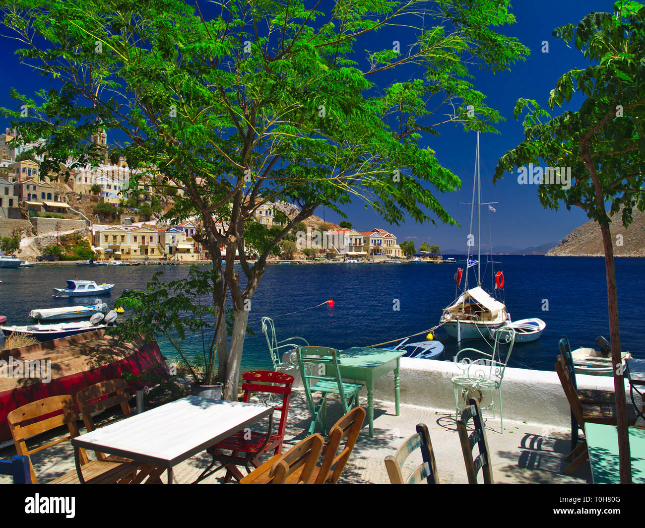Ein Sommer Schuß von der Insel Symi in Griechenland. Blick von einem Café mit Tischen, Sesseln Boote kristallklaren Meer und den bunten Häusern. Stockfoto