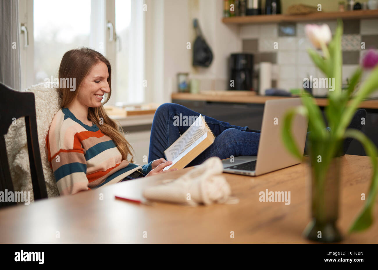 Schöne blonde Frau an einem Tisch sitzen, ein Buch zu lesen, lächelnd, Laptop und häkeln Utensilien auf dem Tisch Stockfoto