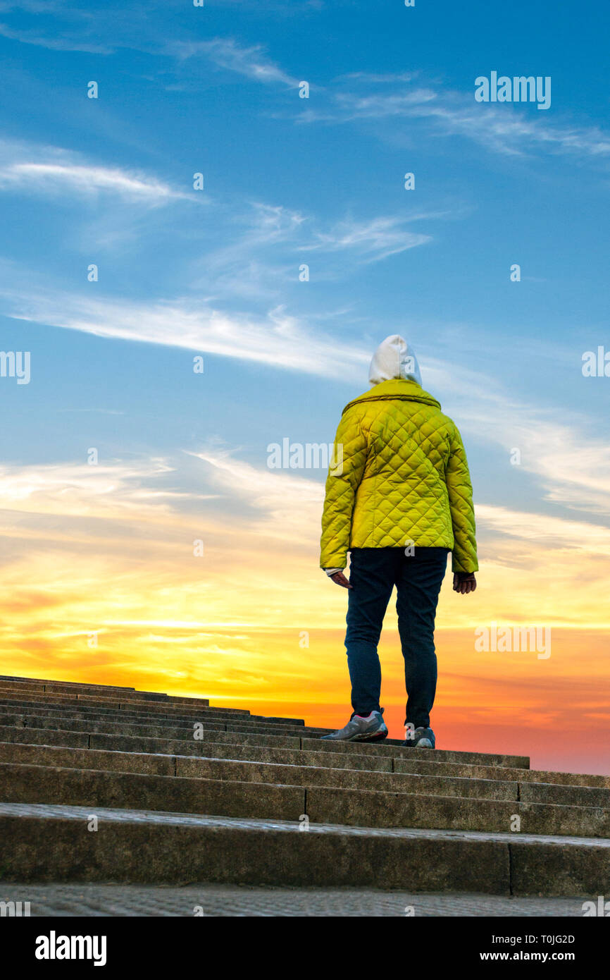 Low Angle Rückansicht Bild der Frau in einem gelben gefütterte Jacke und Kapuze, stehen an der Spitze der Steintreppe in der Morgendämmerung oder Sonnenuntergang Himmel suchen Stockfoto