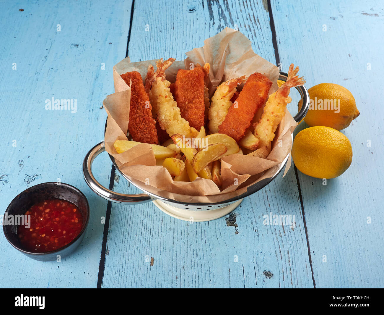 Gebratene Fischstäbchen, Tempura Garnelen und Kartoffelecken in einem Sieb, mit Zitronen und sriracha Soße, auf blauen Hintergrund Holz Stockfoto