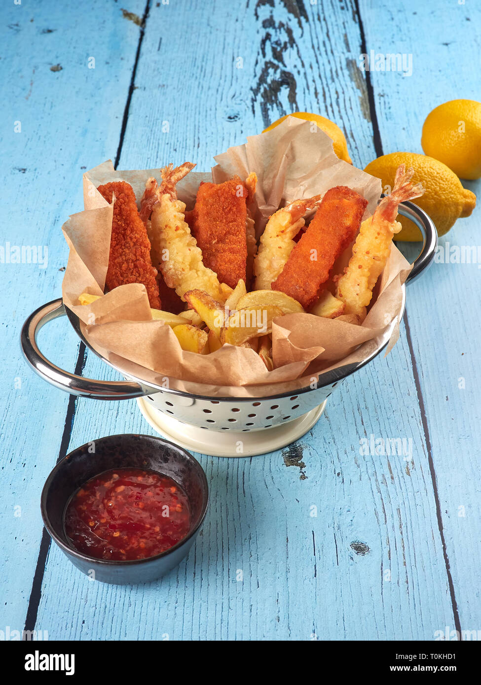 Gebratene Fischstäbchen, Tempura Garnelen und Kartoffelecken in einem Sieb, mit Zitronen und sriracha Soße, auf blauen Hintergrund Holz Stockfoto