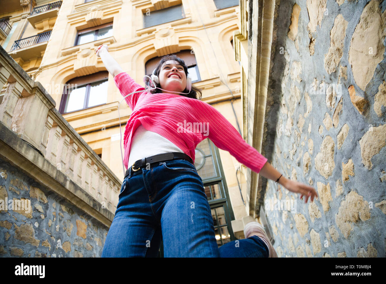 Glückliche junge Frau in rosa Pullover Musik hören und Springen in der Straße. Low Angle View Stockfoto