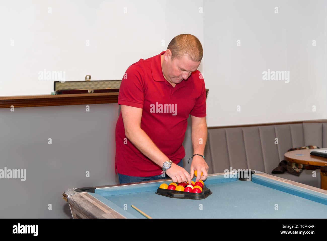 Ein Mann mittleren Alters in einem roten Hemd legt die Billardbälle vorsichtig auf einen Bartisch und bereitet sich auf ein Spiel in einer ungezwungenen Pub-Umgebung vor. Stockfoto