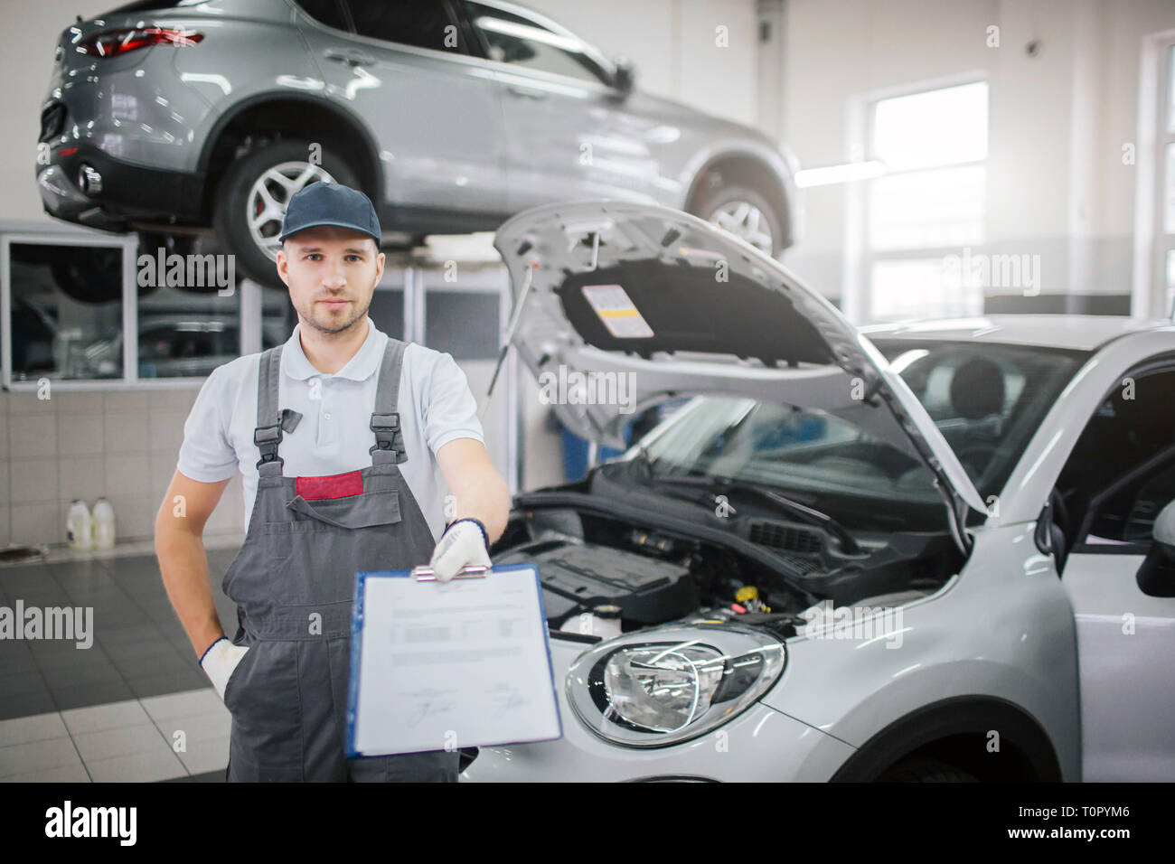 Schön und selbstbewussten jungen Mensch steht im Auto und schaut auf Kamera. Er hält Plastik Ordner mit Dokumenten auf. Eine Hand in der Tasche. Körper des Auto ist op Stockfoto