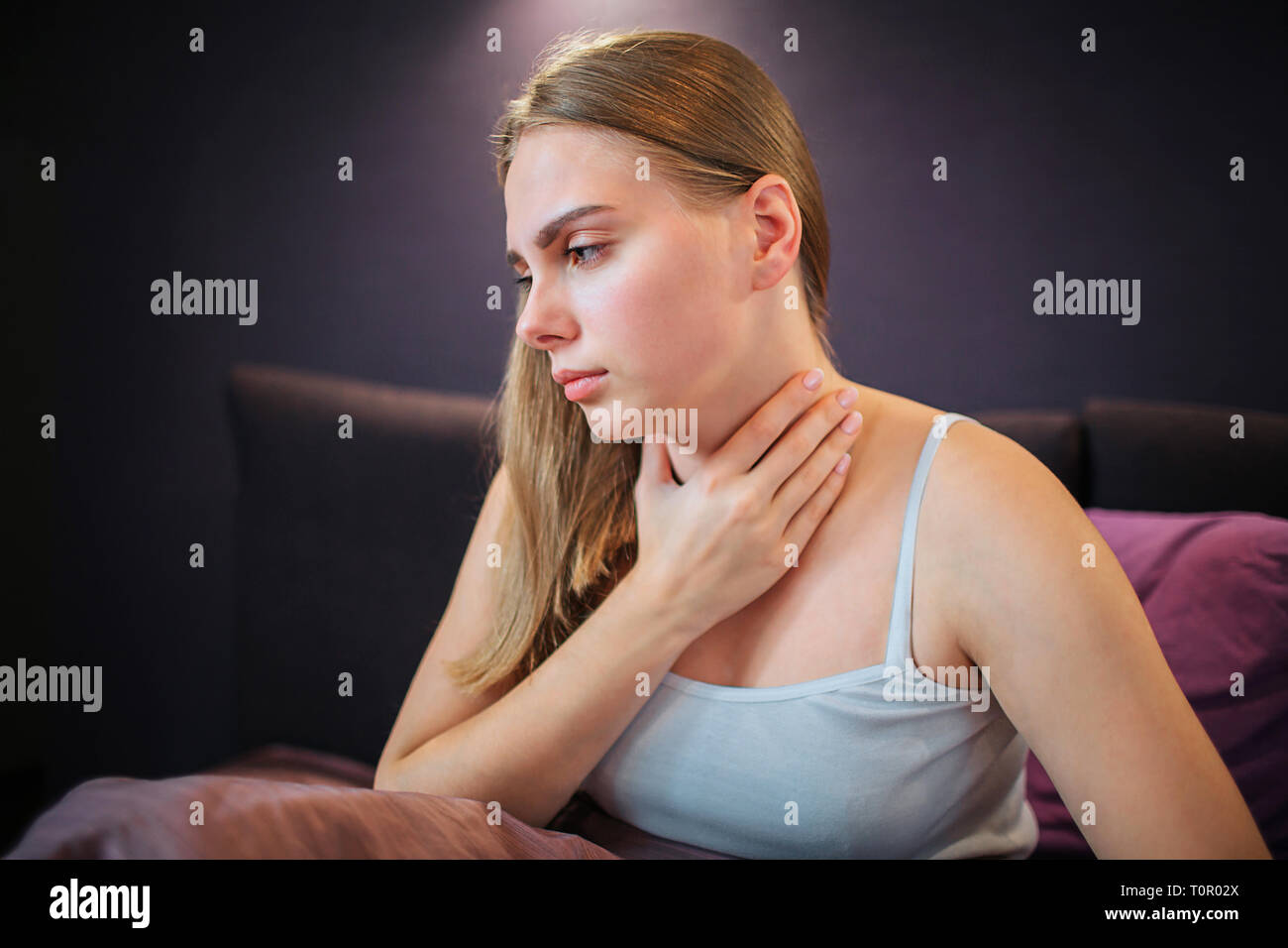 Sorgen junge Frau sitzt auf dem Bett und schaut nach unten. Sie hält die Hand auf den Hals. Frau fühlt es den Schmerz. Stockfoto