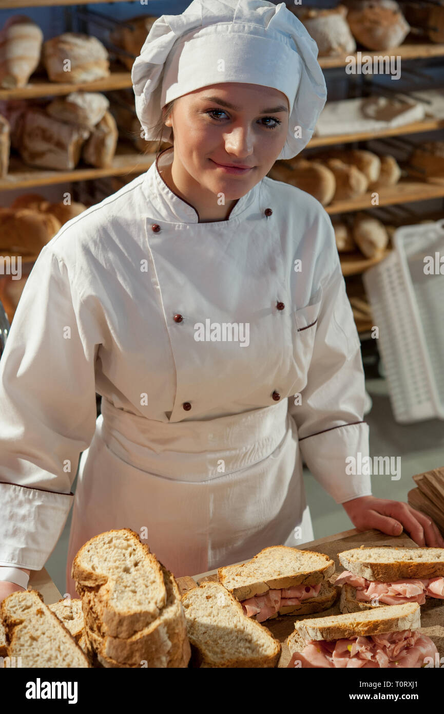 Lächelnden jungen Verkäuferin in der Bäckerei Schreibtisch bereitet Mortadella Bologna Sandwiches. Regale voll Brot in den Hintergrund. Stockfoto