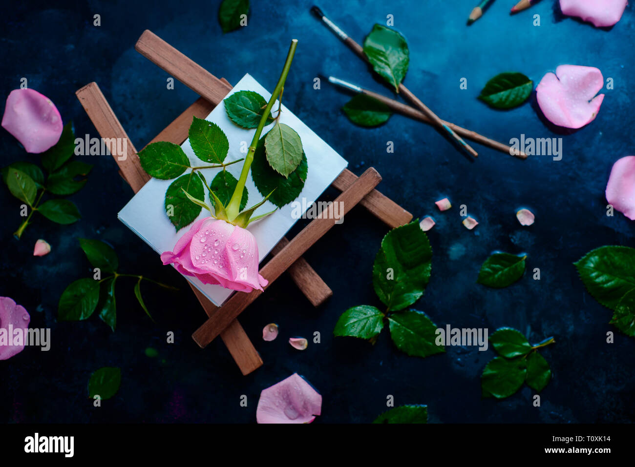 Rose Blume auf einem Maler Staffelei flach. Werkzeuge, Pinsel und Bleistift auf einem nassen regnerischen Hintergrund mit kopieren. Natur und Kunst Konzept Stockfoto