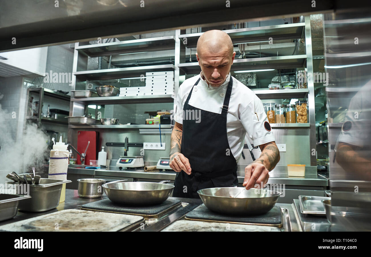 Vertrauen in alles. Schweren männlichen Chef mit mehreren Tattoos auf seinem Arme Braten Zutaten für seine Speise in einem Restaurant Küche. Kochvorgang. Essen Blogger Stockfoto