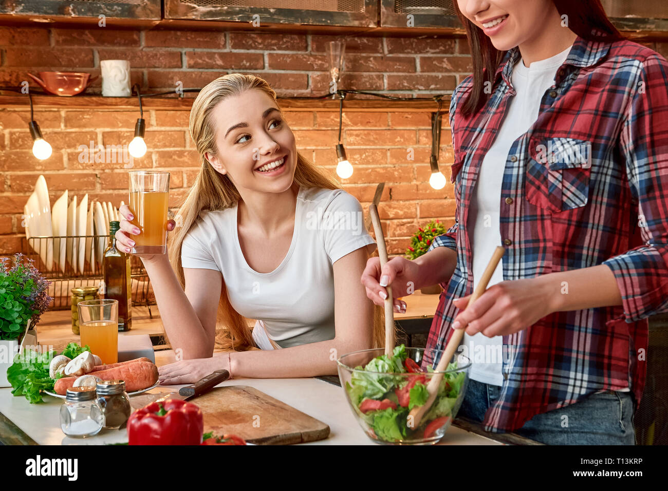 Porträt von zwei attraktive Frauen, Spaß zu haben, während der Vorbereitung Gemüse Salat. Sie sind voll in den Prozess einbezogen. Blonde Mädchen im weißen T-Shirt betrachtet ihren Freund mit einem Lächeln. Ihr dunkelhaarigen Freund in kariertem Hemd ist das Mischen von gehacktes Gemüse in eine Schüssel geben. Stockfoto