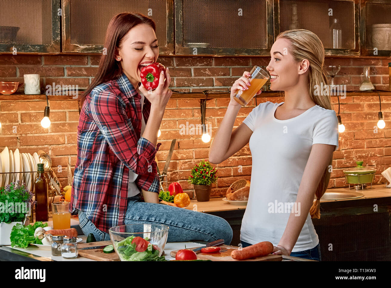 Porträt von zwei attraktive Frauen, Spaß zu haben, während der Vorbereitung Salat. Sie sind voll in den Prozess einbezogen. Dunkelhaarige in kariertem Hemd sitzen auf die Tischplatte, sie wird ein Pfeffer zu beißen. Ihre blonden Freund im weißen T-Shirt Getränke frischer Saft und betrachtet ihre Stockfoto