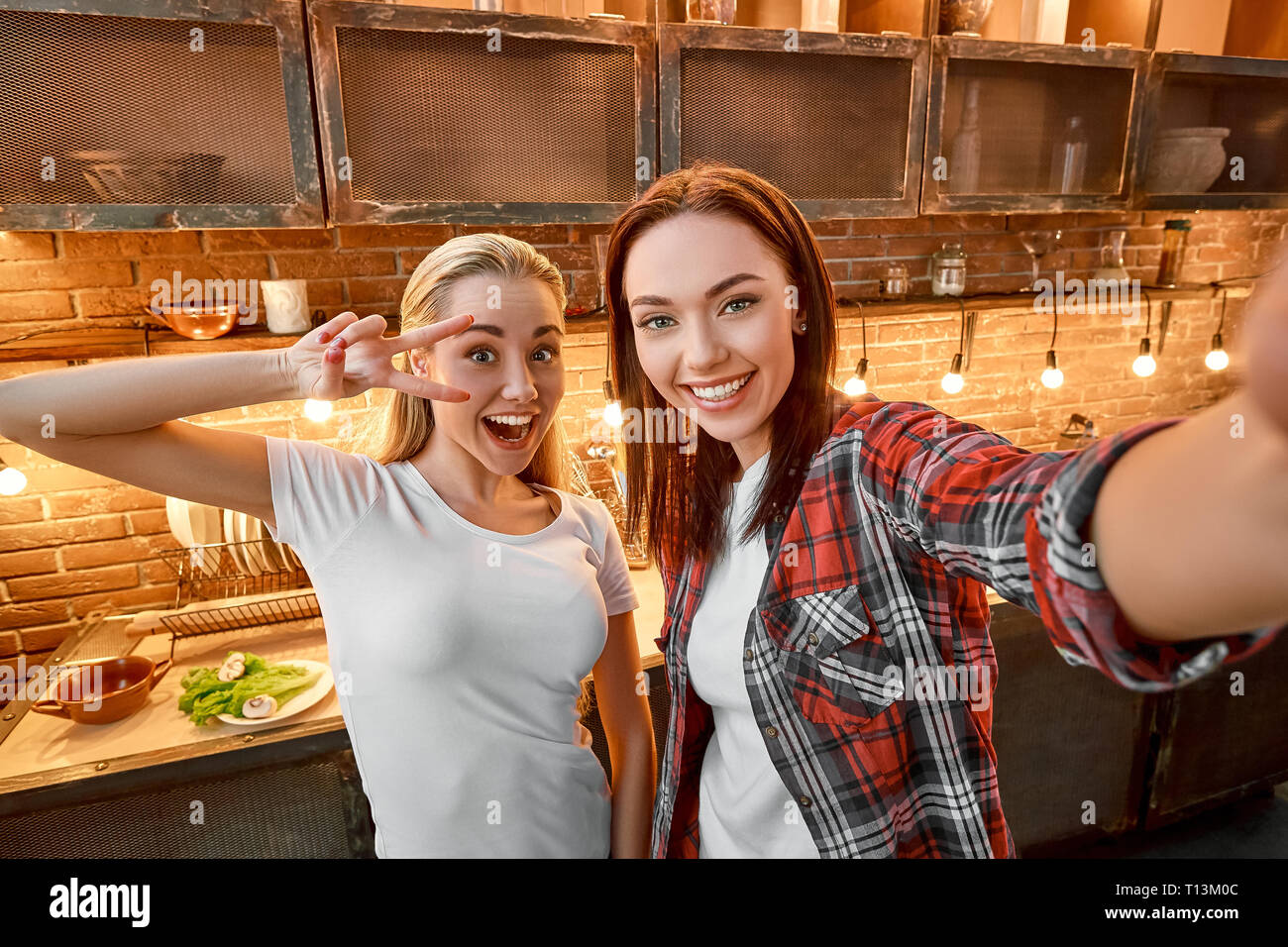 Selfie von zwei fröhlichen Frauen, Spaß haben, während der Vorbereitung ein Gericht. Dunkelhaarige Mädchen mit kariertem Hemd ist ein Foto, während ihre blonde Freundin für Sie stellt. Sie zeigt V-Schild in der Nähe ihre Augen und lächelte. Stockfoto