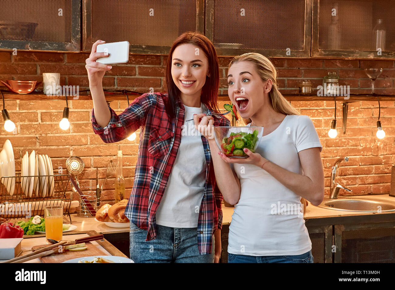 Porträt von zwei fröhlichen Frauen, Spaß haben, während der Vorbereitung Gemüse Salat. Dunkelhaarige Mädchen mit kariertem Hemd nimmt selfie, während ihre blonde Freundin im weißen T-Shirt einem gekochten Salat hält und posiert für ein Foto Stockfoto