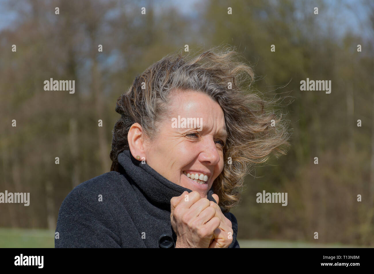 Lächelnde Frau mittleren Alters an einem windigen Tag auf dem Lande, kuschelte sich in eine warme Jacke, wie ihr Haar weht im Wind frei Stockfoto