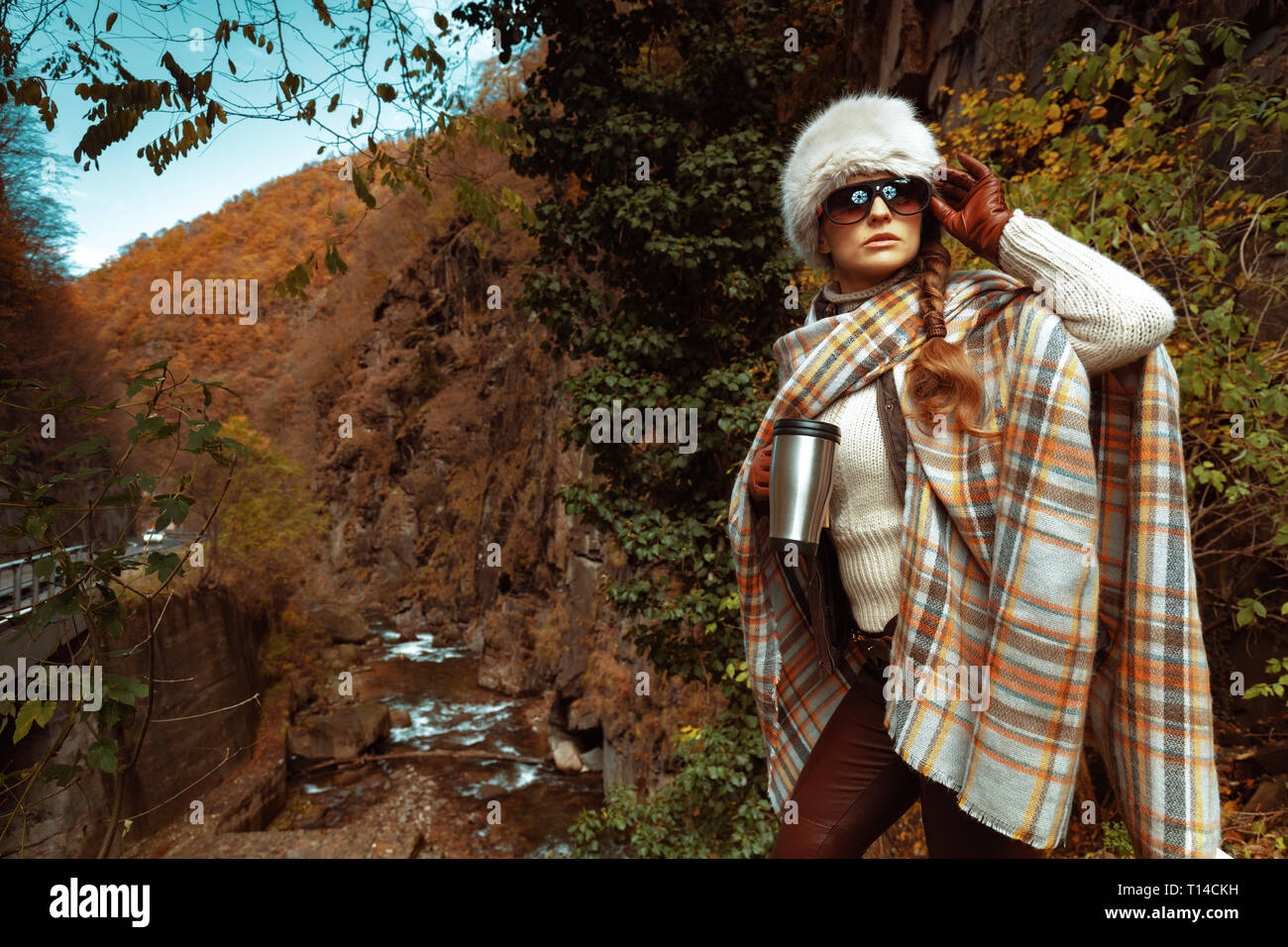 Junge touristische Frau in Poncho und Sonnenbrille mit Edelstahl thermos Travel Mug Blick in die Ferne in den Wald. Stockfoto
