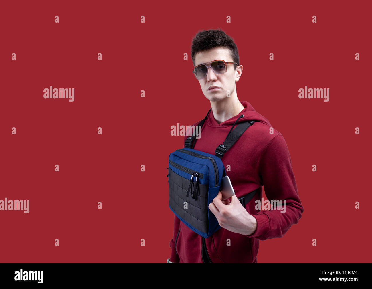 Junger hübscher Kerl in Sonnenbrille und roter Kapuze mit Brust rig Bag posiert im Studio auf einem roten Hintergrund Modell schießen Stockfoto