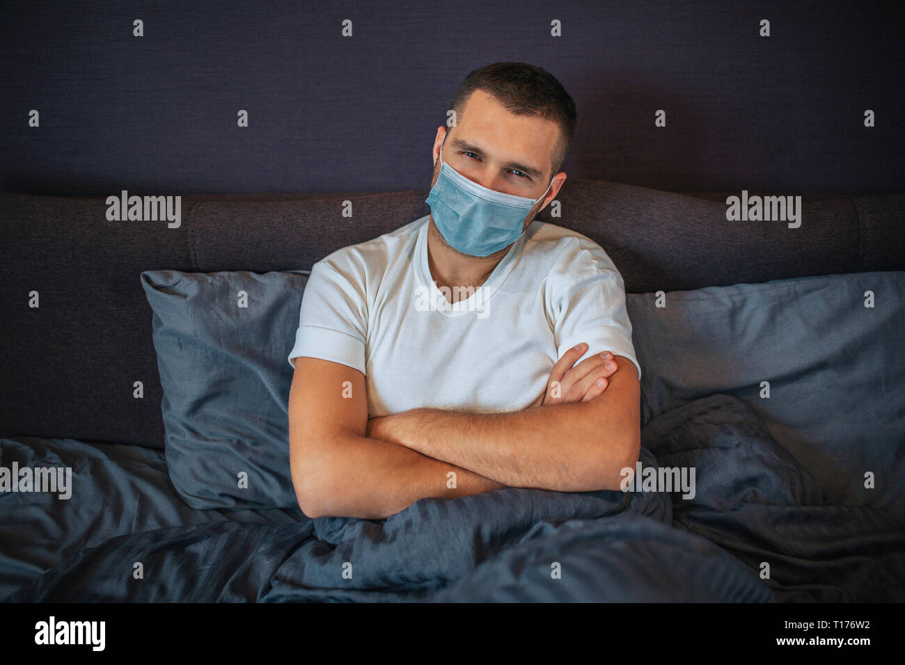 Ernsthafte junge Mann trägt Schutz Maske. Er sitzt auf dem Bett und schaut auf Kamera. Kerl hält die Hände gekreuzt. Er ist sehr ernst. Stockfoto
