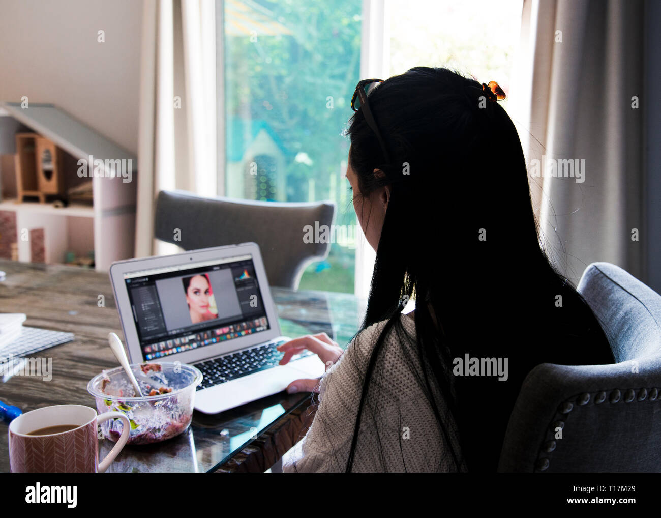 Junge Frau, die an einem Schreibtisch auf ihrem Laptop sitzt und von der Seite aus einen Blick auf das Haus hat. Stockfoto