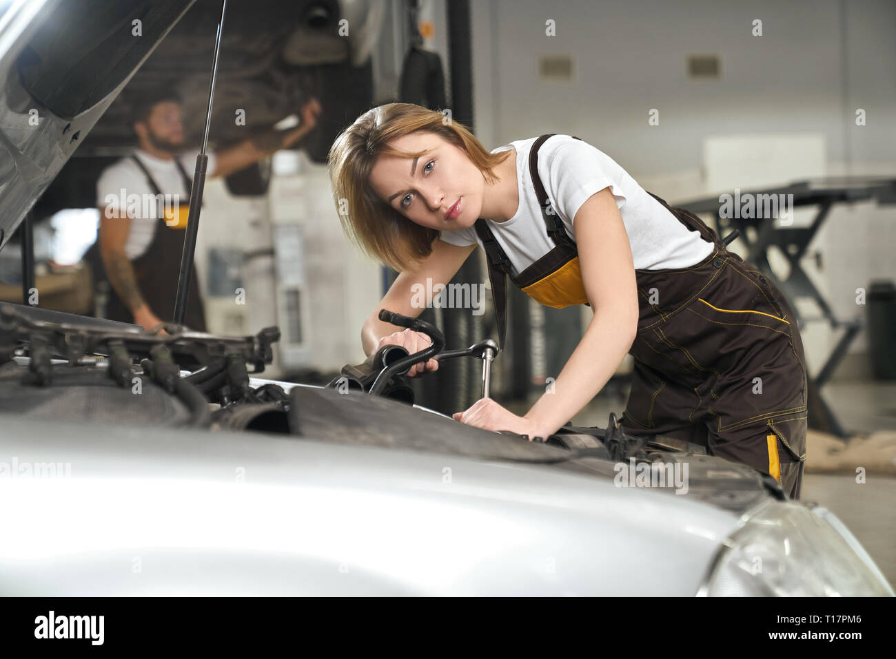 Mechaniker Mädchen tragen in Overalls und weißes T-Shirt Auto reparieren. Hübsche junge Frau lehnte sich auf dem Auto, Instrument und Kamera, posiert in Autoservice. Stockfoto