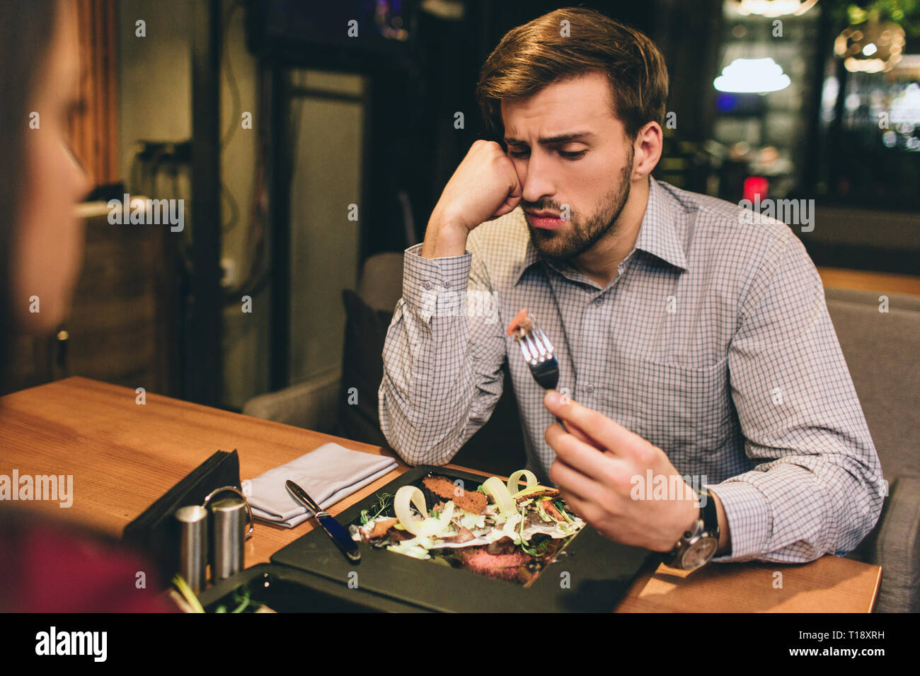 Foto der Kerl sitzt zusammen mit seiner Freundin und Essen einige Lebensmittel, die Sie bestellt haben. Der Mensch hat etwas Fleisch auf dem Teller gefunden. Er ist auf der Suche nach Stockfoto