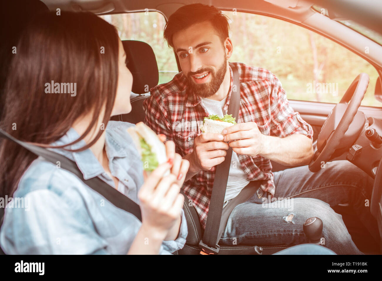 Schön und gut aussehendes Paar ist sitzen zusammen im Auto und miteinander zu suchen. Sie sind mit dem Sicherheitsgurt gesichert. Die Menschen halten die Sandwiches. T Stockfoto