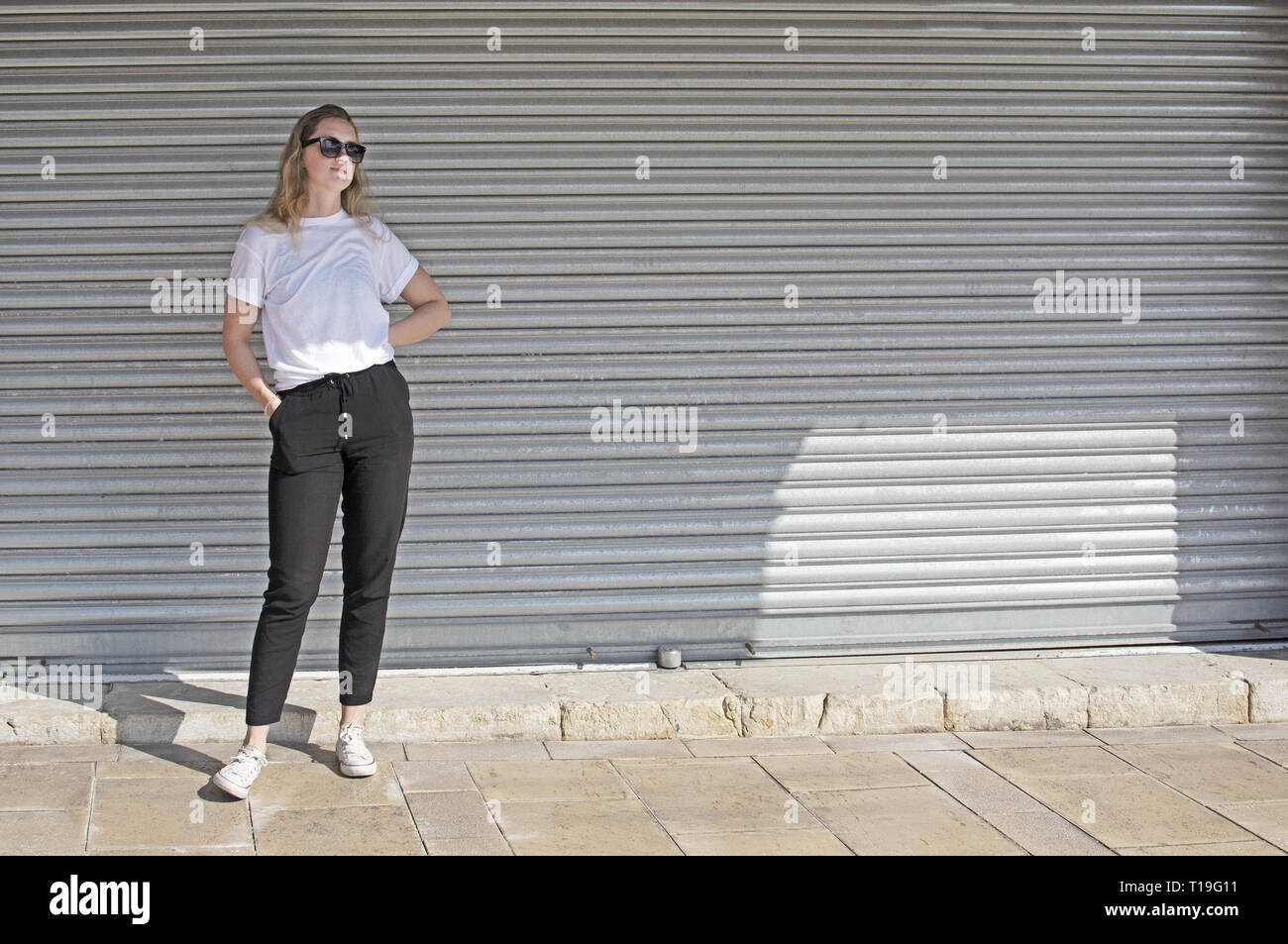 Voller Körper Foto der jungen Casual sportlich gekleidet blonde Frau mit Sonnenbrille im weißen T-Shirt gegen Wellblech wall street style Stockfoto