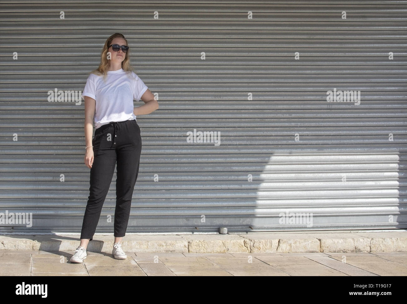 Voller Körper Foto der jungen Casual sportlich gekleidet blonde Frau mit Sonnenbrille im weißen T-Shirt gegen Wellblech wall street style Stockfoto